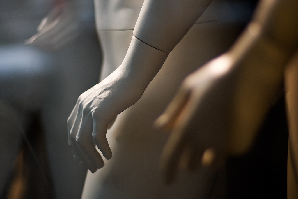 A white plastic mannequin hand lit from the upper left side of the image in a store window. Two other palms are visible out of focus in the foreground and background.